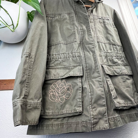 close up angled view of a military green cotton parka with many pockets, the right hip pocket has an outline of a peony hand embroidered in peach on it