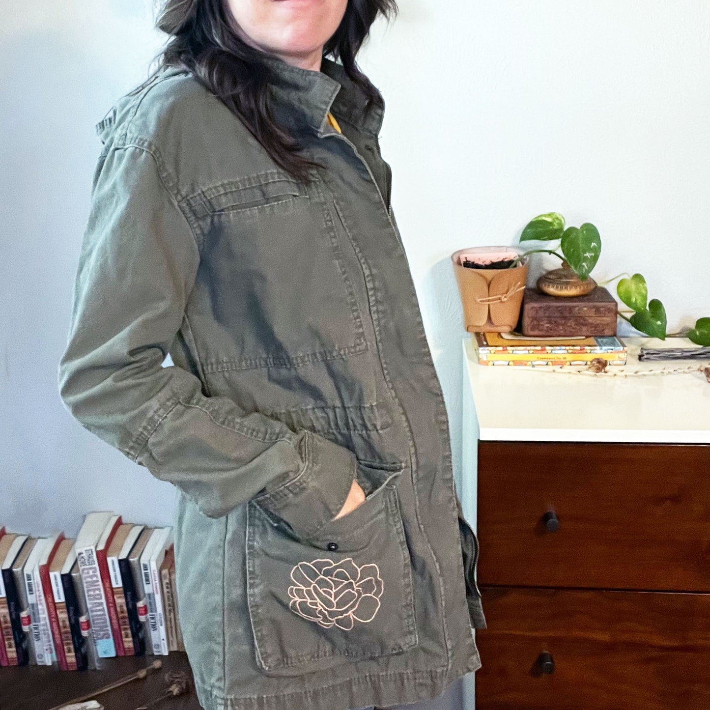 side view of a woman wearing a military green cotton parka with many pockets and a hood, the right hip pocket has an outline of a peony hand embroidered in peach on it, standing in front of a white and wood dresser with a plant on it and a table with books on it