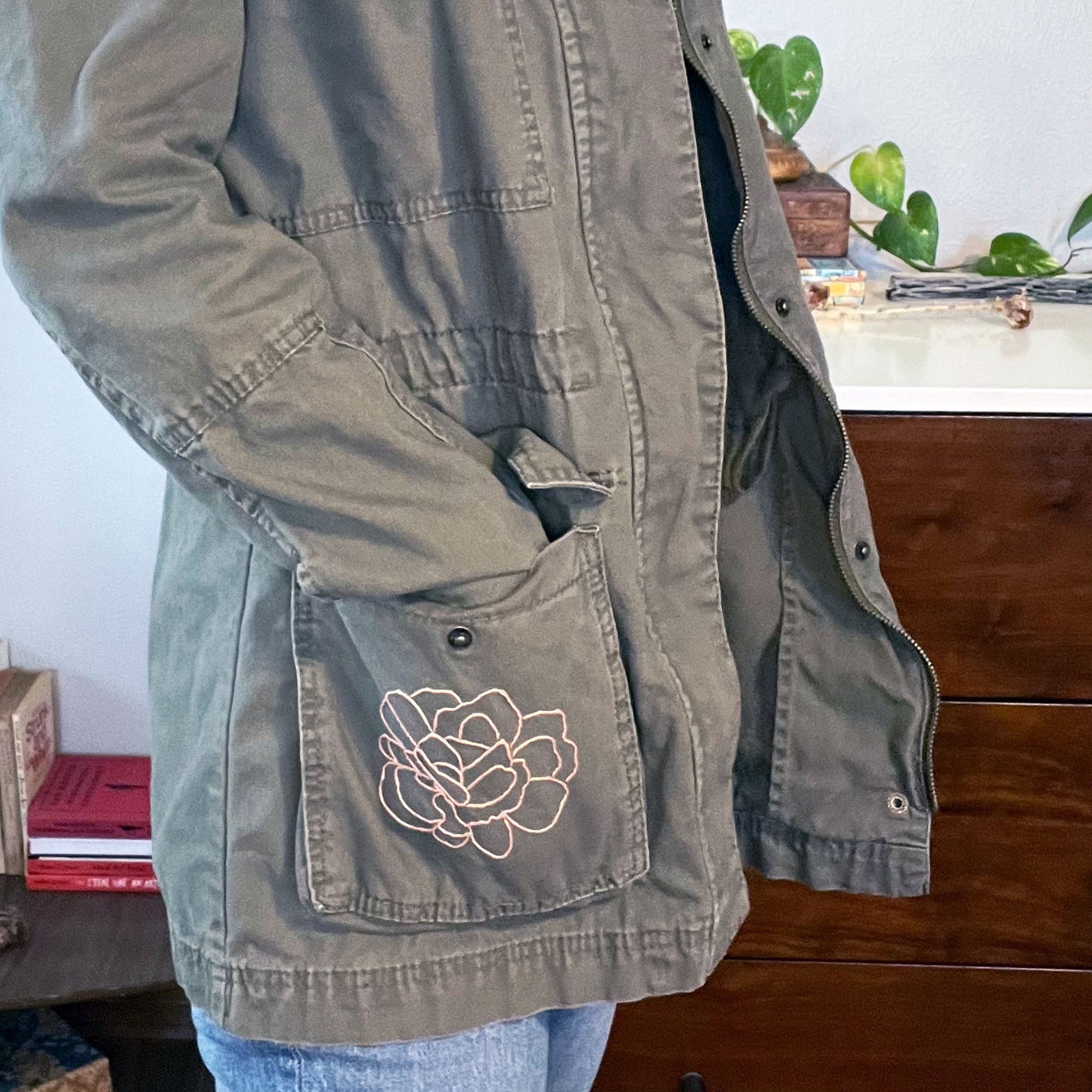 close up view of side view of a woman wearing a military green cotton parka with many pockets and a hood, the right hip pocket has an outline of a peony hand embroidered in peach on it, standing in front of a white and wood dresser with a plant on it and a table with books on it