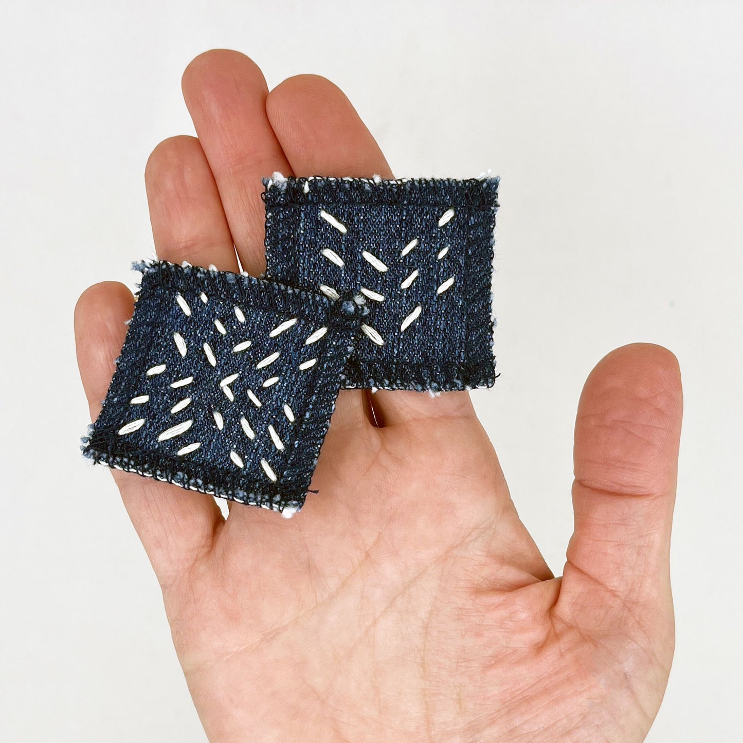 a hand palm up holding two square denim patches with running stitches in ecru of three chevron lines and a repeating x pattern on a white background