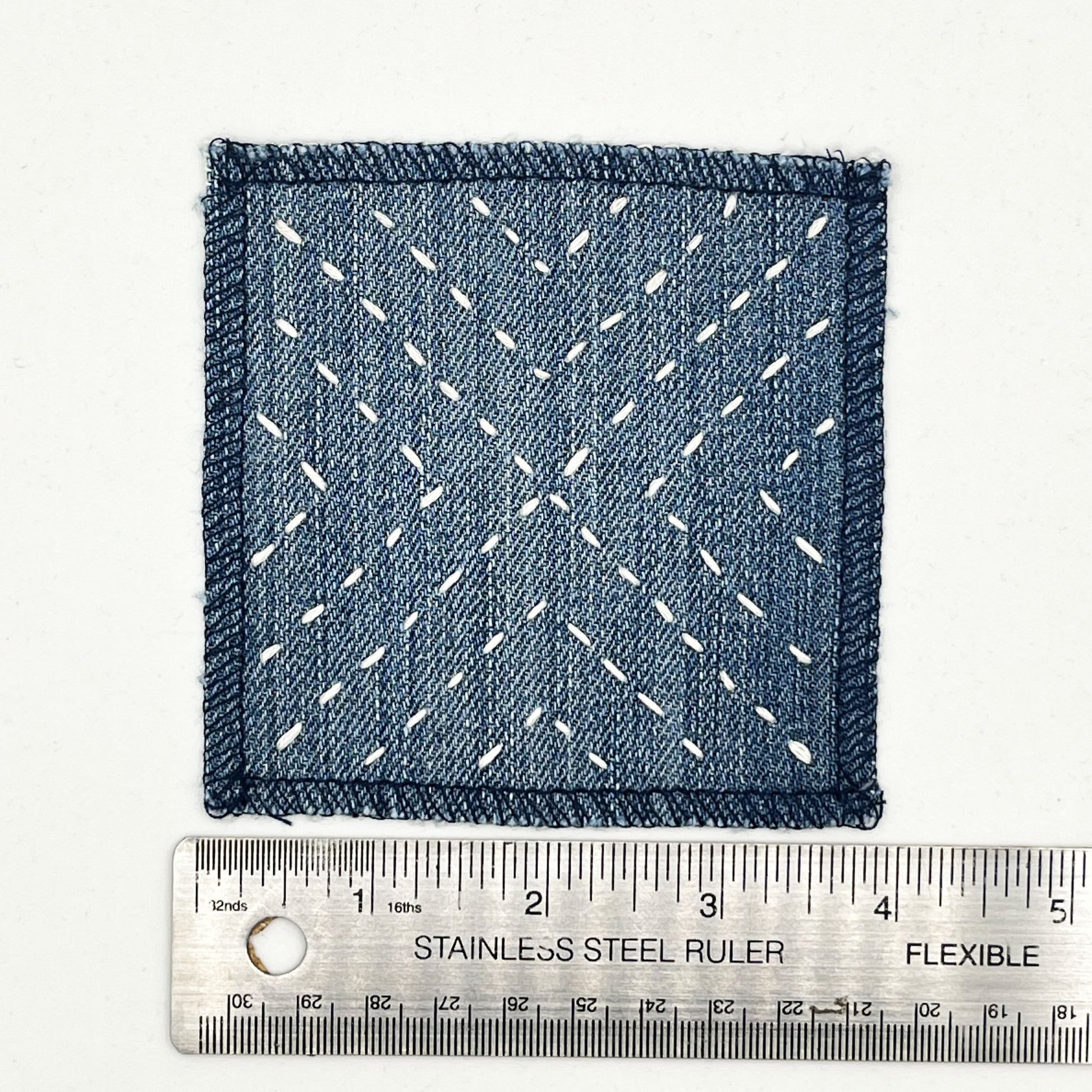 a square patch made out of denim, handstitched with ivory running stitches in a radiating X pattern, with overlocked edges, next to a metal ruler to show a width of 4 inches, on a white background