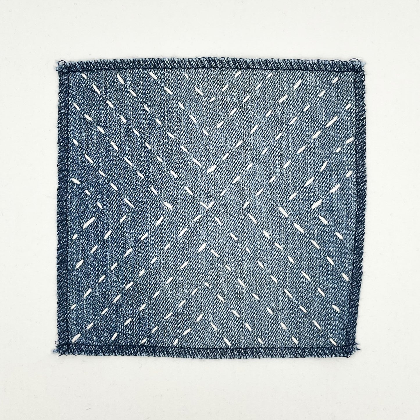 a square patch made out of denim, handstitched with ivory running stitches in a radiating X pattern, with overlocked edges, on a white background