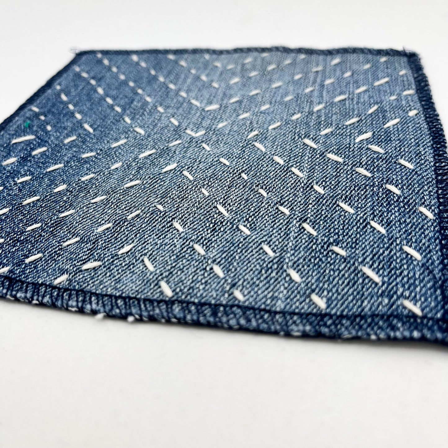close up angled view of a square patch made out of denim, handstitched with ivory running stitches in a radiating X pattern, with overlocked edges, on a white background