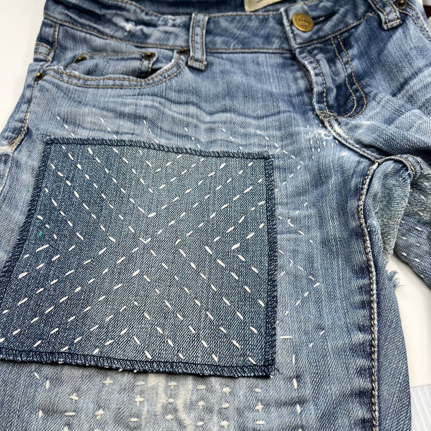 close up angled view of a square patch made out of denim, handstitched with ivory running stitches in a radiating X pattern, with overlocked edges, on a pair of jeans with other visible mends