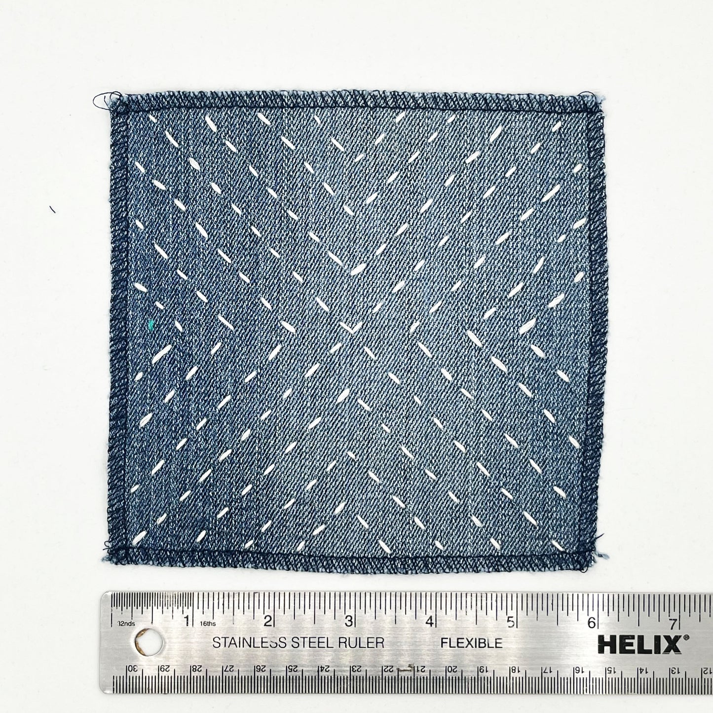 a square patch made out of denim, handstitched with ivory running stitches in a radiating X pattern, with overlocked edges, next to a metal ruler to show a width of 6 inches, on a white background