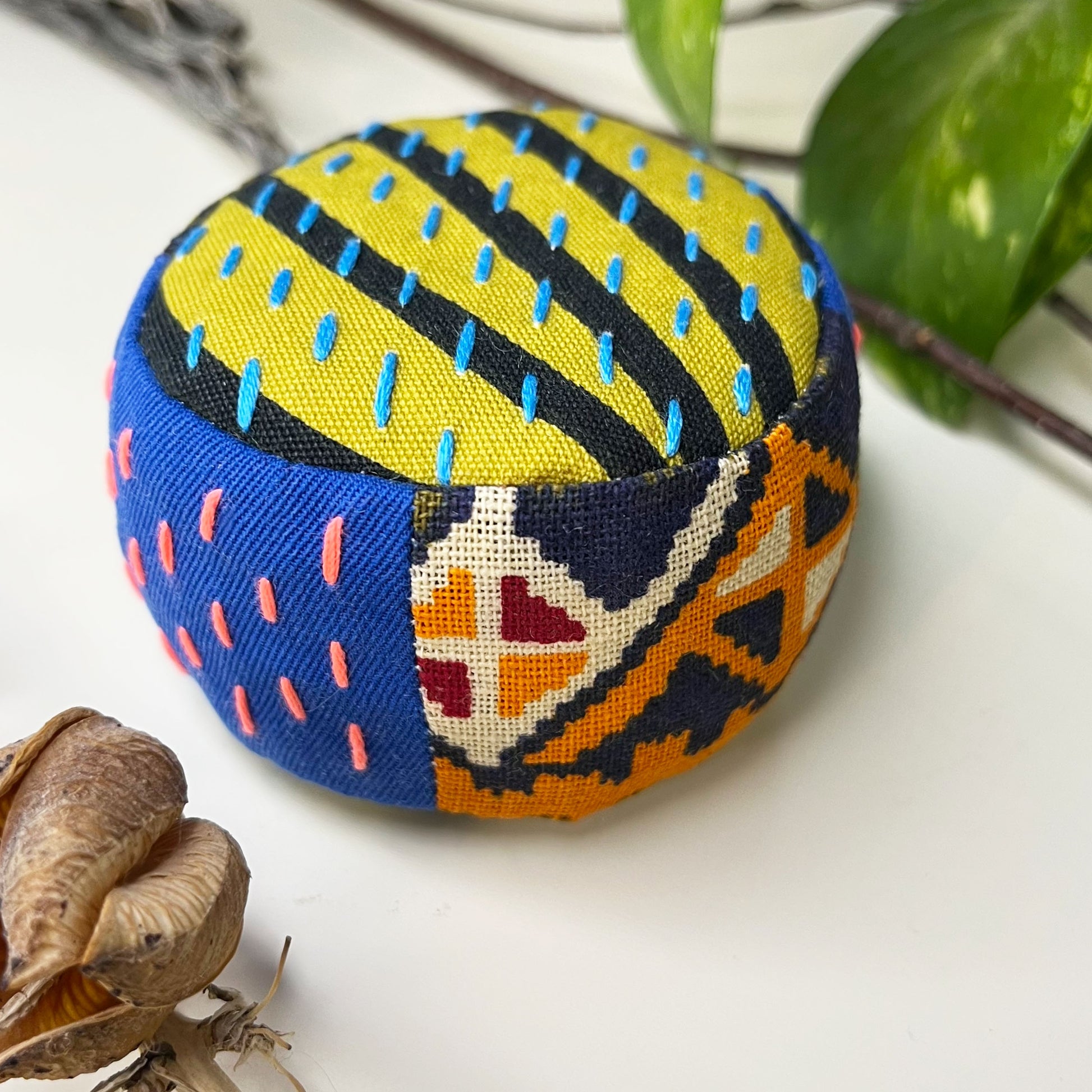 a round pincushion made from brightly colored patterned and solid fabric with contrasting and also brightly colored sashiko stitches on some side panels and the top