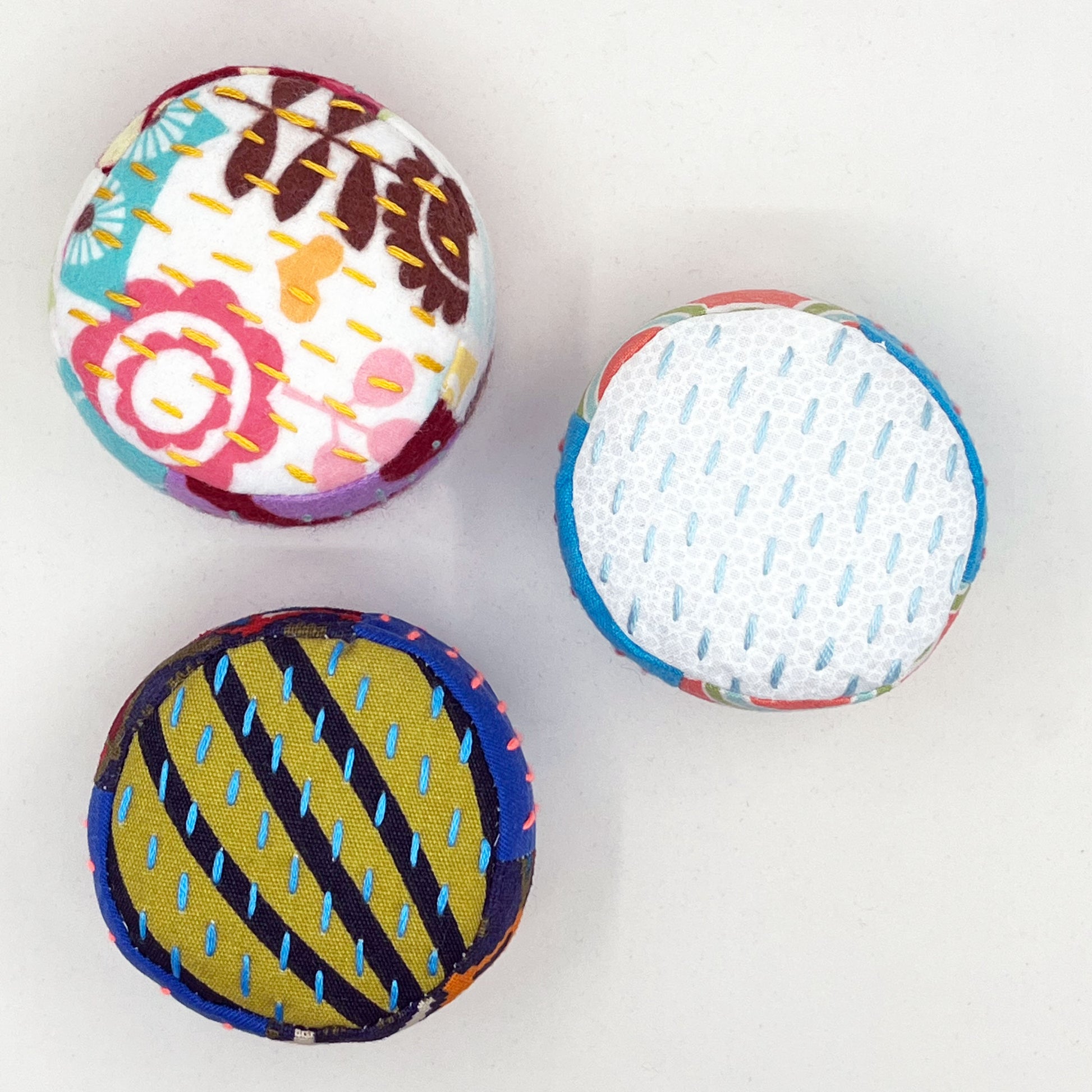 view from above of a group of 3 colorful pincushions on a white counter, with sashiko stitching in contrasting colors