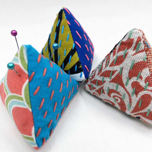 three pyramid shaped pincushions, made from different combinations of colorful fabric, some with sashiko stitching on contrasting colors, on a white background