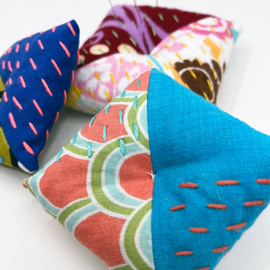 three square shaped pincushions, made from different combinations of colorful fabric with diagonal seams, with sashiko stitching in contrasting colors, on a white background