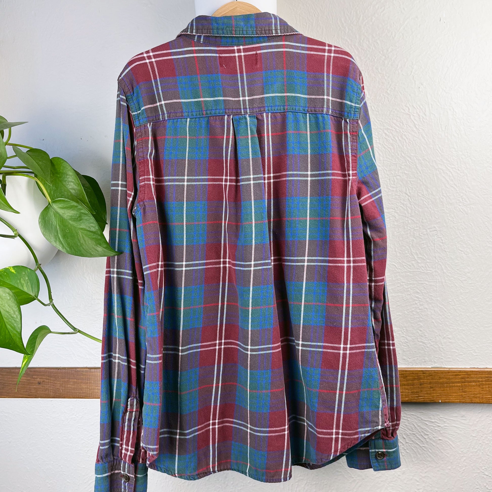 back view of a maroon green and blue plaid flannel on a hanger next to a wall with a plant next to it