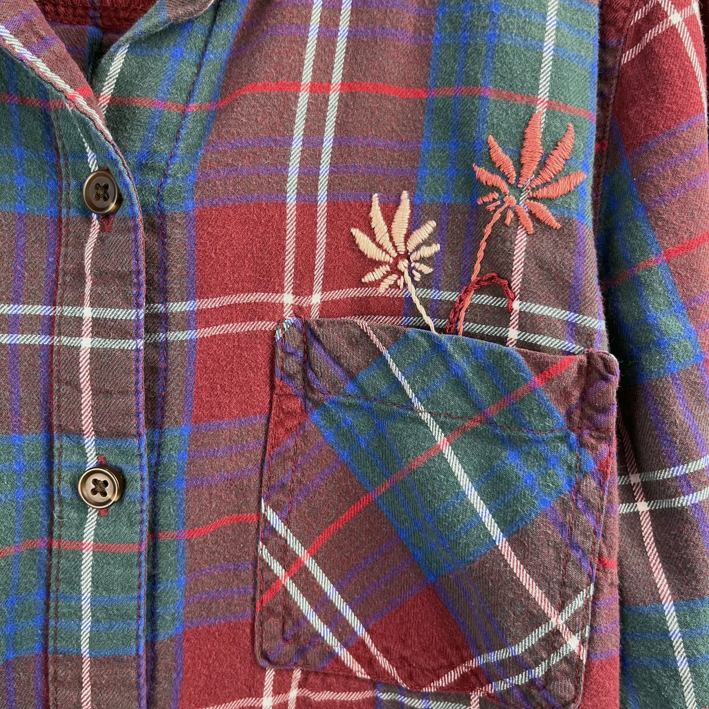 close up view of a maroon green and blue plaid flannel with two hand embroidered mauve flowers coming out of the chest pocket