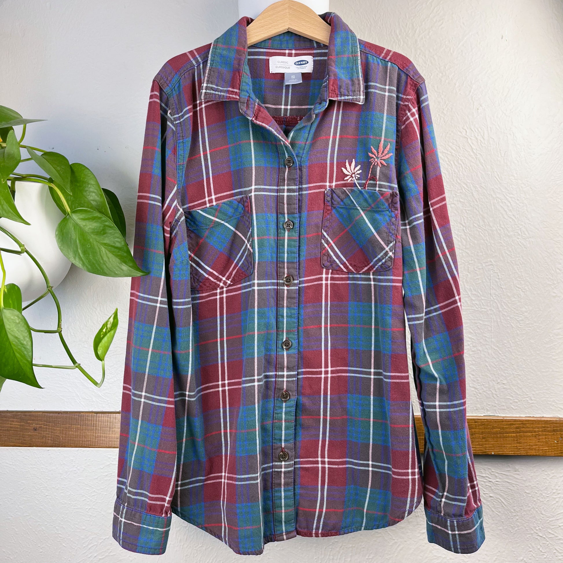 a maroon green and blue plaid flannel with two hand embroidered mauve flowers coming out of the chest pocket on a hanger next to a wall with a plant next to it