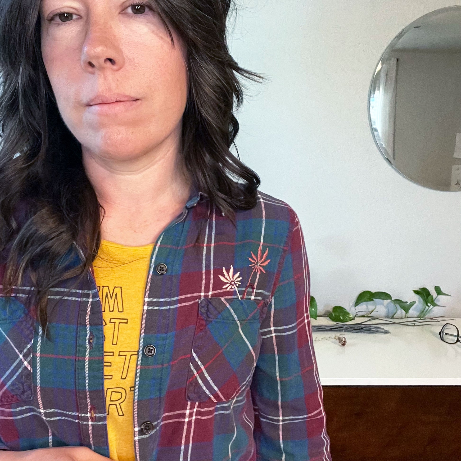 a woman with curly brown hair wearing a maroon green and blue plaid flannel with two hand embroidered mauve flowers coming out of the chest pocket, standing in front of a white dresser with a plant vine on it and a circle mirror on the wall