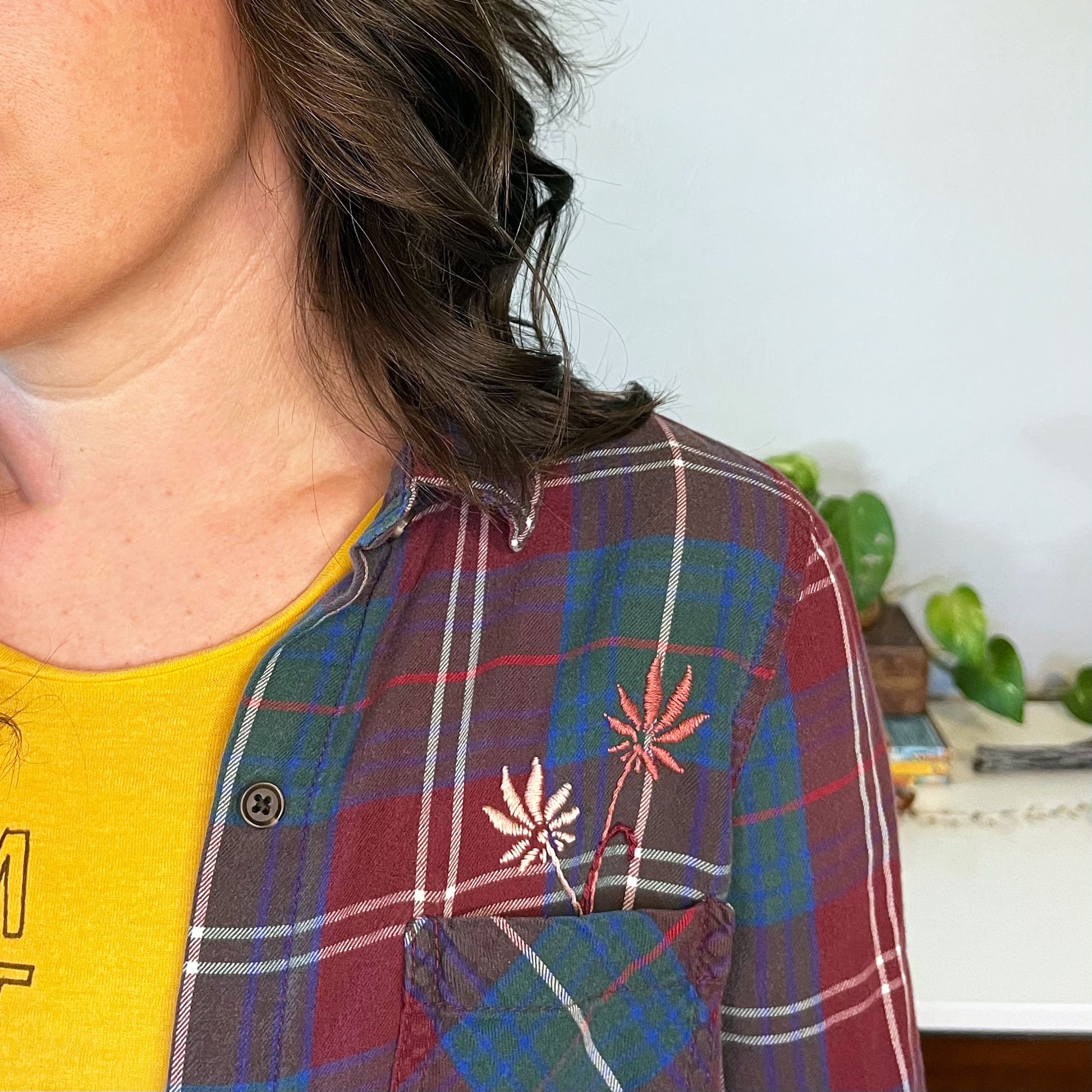 close up view of a woman wearing a maroon green and blue plaid flannel with two hand embroidered mauve flowers coming out of the chest pocket