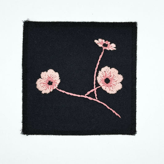 a black colored square patch, hand embroidered with three poppies on a stem, in shades of pink, with overlocked edges, on a white background