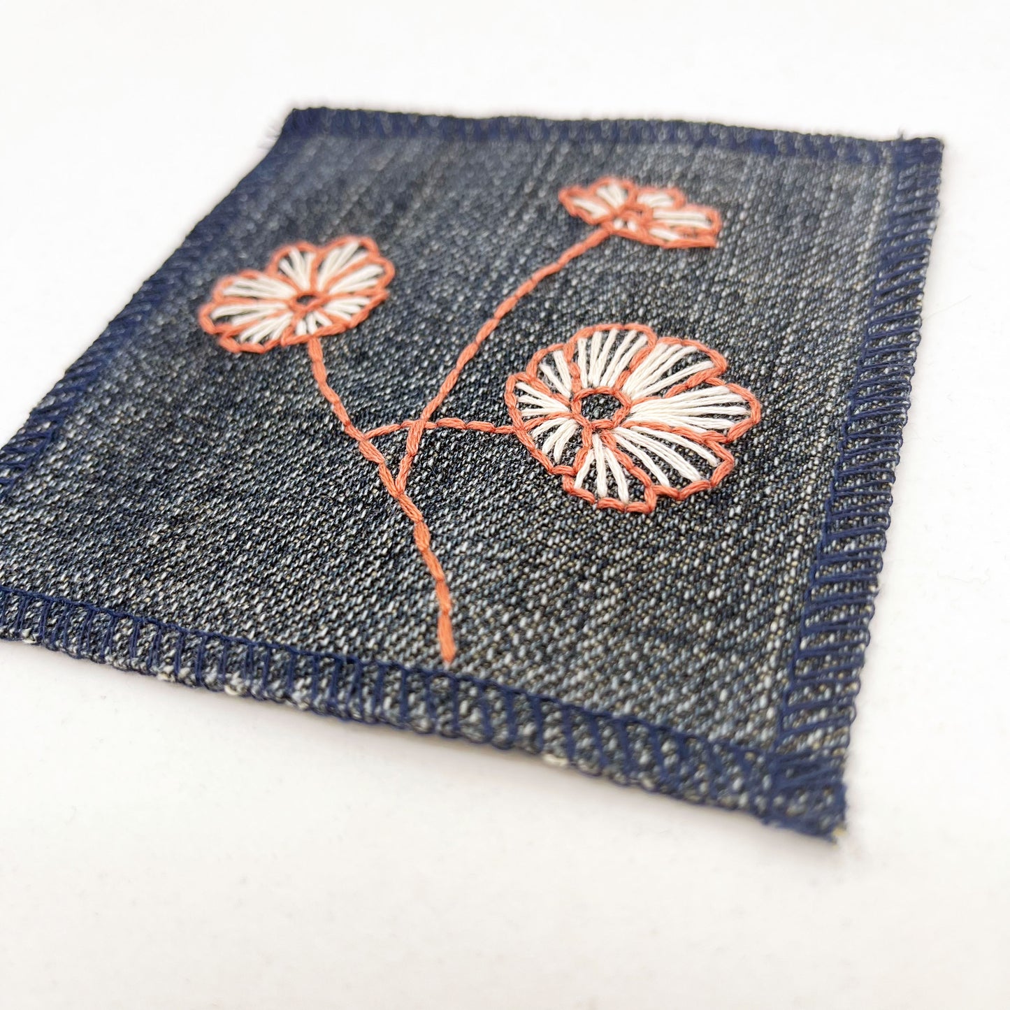 close up angled view of a square denim patch with three poppies hand embroidered on it, the outline of the petals and stem in clay colored thread and the fill lines in peach