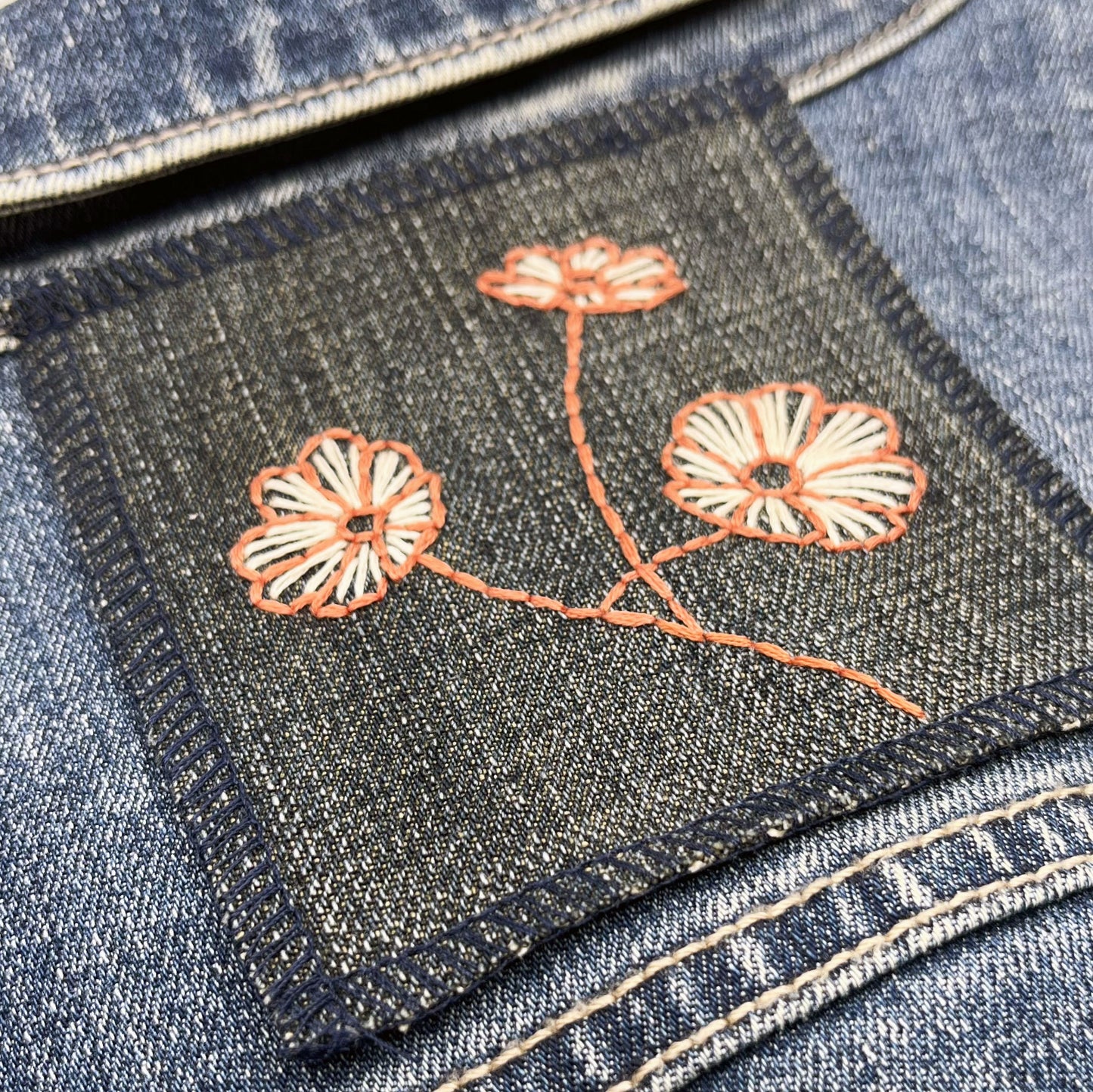 a square denim patch with three poppies hand embroidered on it, the outline of the petals and stem in clay colored thread and the fill lines in peach on a denim jacket