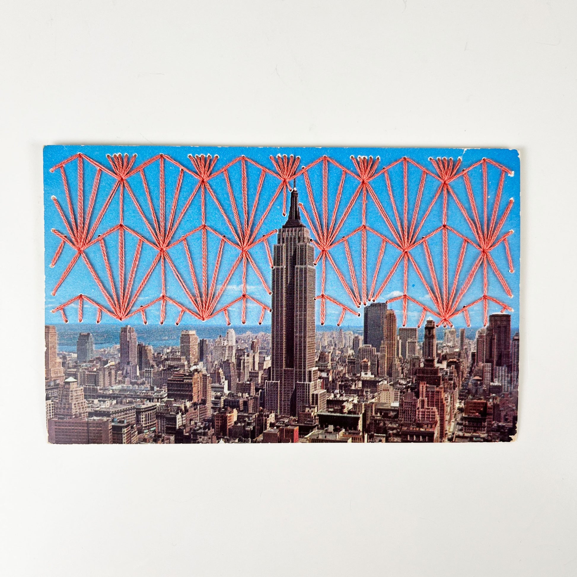 an old postcard of the Empire State Building, with a hand embroidered Art Deco design in coral stitched in the background on the sky only, the postcard is sitting on a white background