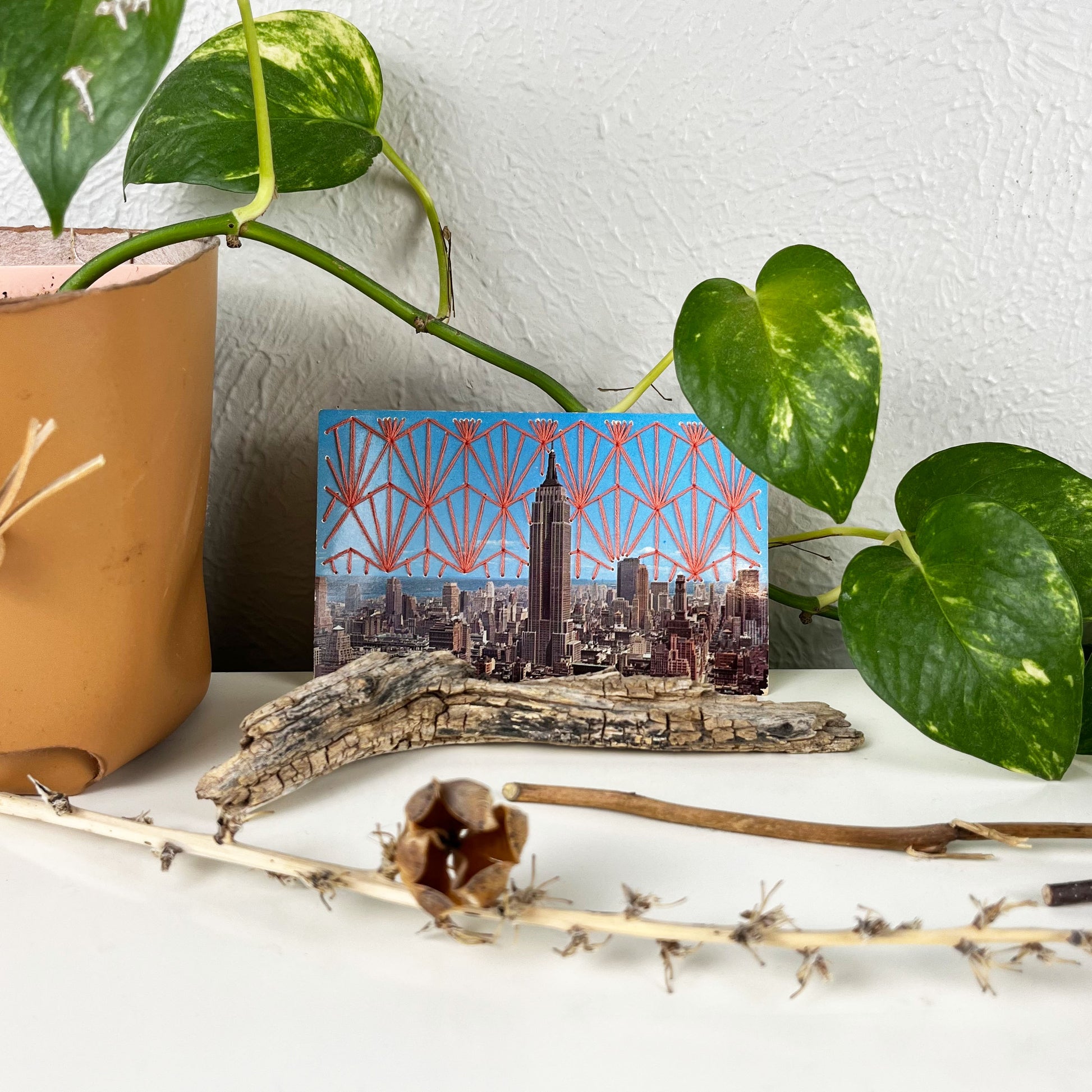 an old postcard of the Empire State Building, with a hand embroidered Art Deco design in coral stitched in the background on the sky only, the postcard is sitting on a white dresser with a plant vine behind it and sticks and dried flowers in front of it