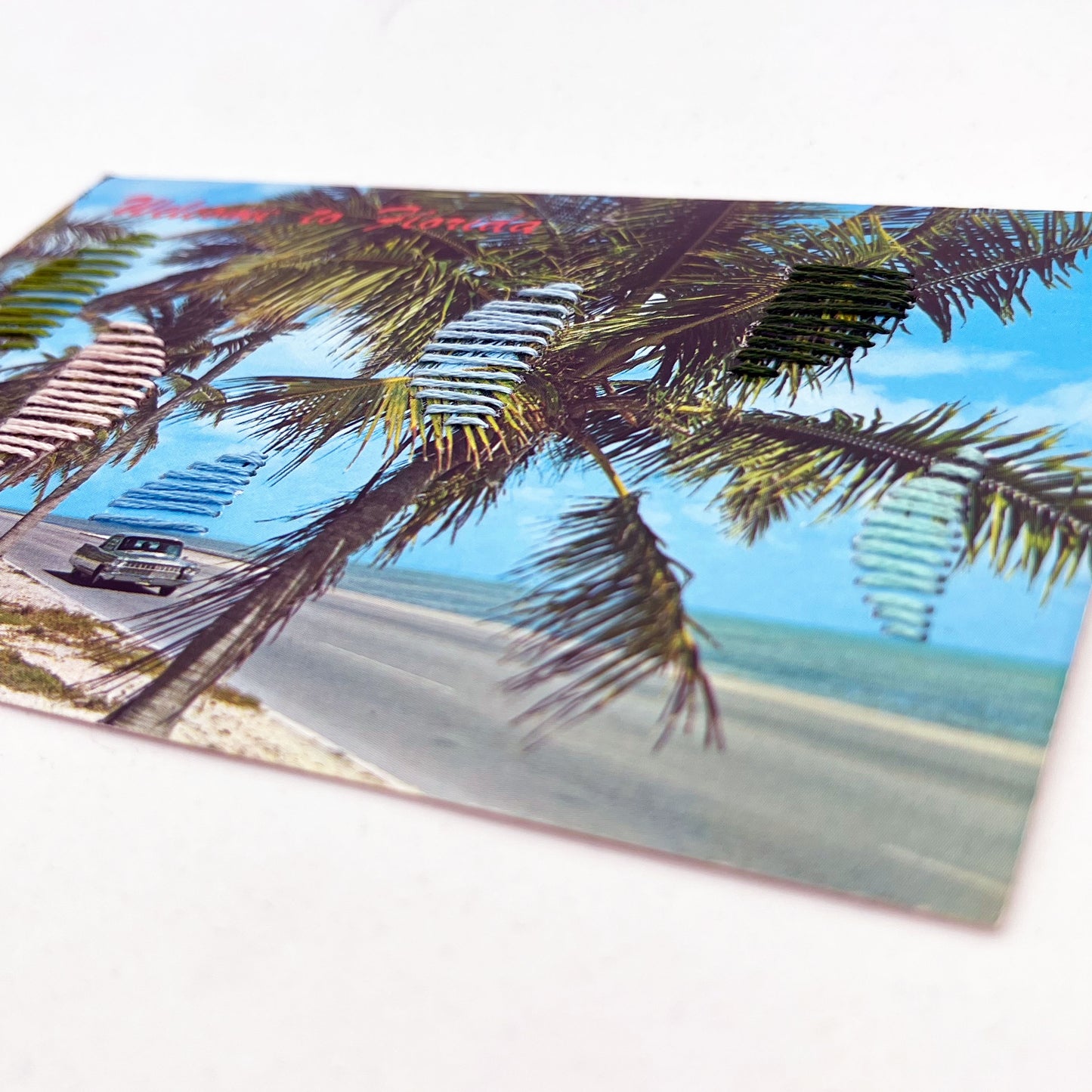 close up angled view of an old postcard of a Florida beach, with hand embroidered half circles randomly placed in greens blues and peaches, on a white surface