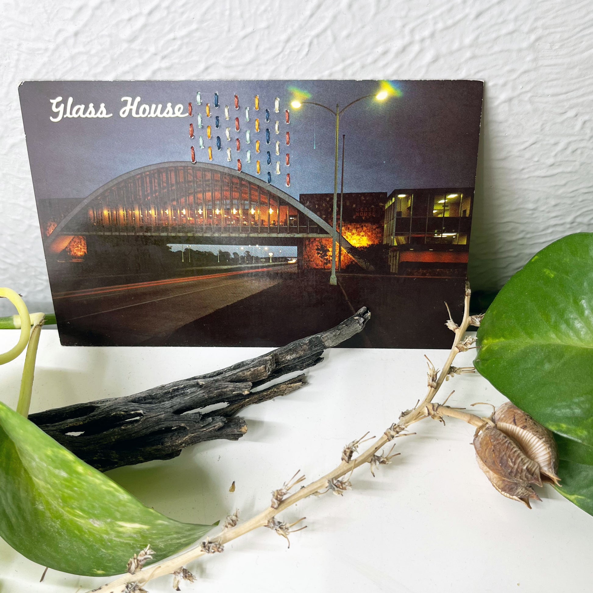 an old postcard of the Glass House rest stop restaurant, with multicolored hand embroidered lines in the sky, it is sitting on a dresser with a plant vine behind it and sticks in front of it