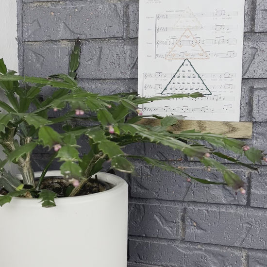 short video clip of Christmas sheet music, hand stitched on with a Christmas tree made from triangles filled with different types of stitches, in green, peach and ivory thread, hanging in a wood magnetic frame on a grey brick wall, with a Christmas cactus in front of it. the video starts far away and zooms in slowly