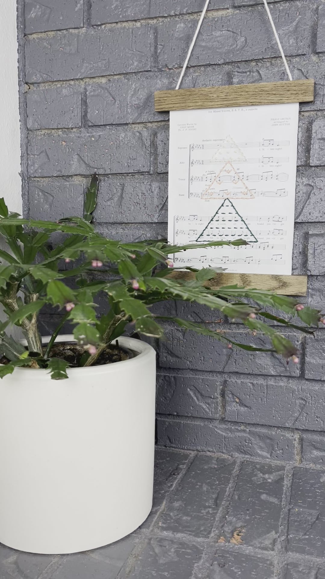 short video clip of Christmas sheet music, hand stitched on with a Christmas tree made from triangles filled with different types of stitches, in green, peach and ivory thread, hanging in a wood magnetic frame on a grey brick wall, with a Christmas cactus in front of it. the video starts far away and zooms in slowly