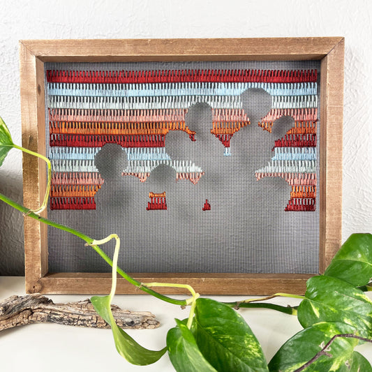 close up view of a piece of window screen hand stitched with rows of maroon rusty orange mint green and light blue stitches with an image in negative space of a prickly pear cactus, in a wood frame, on a white counter, with a pothos vine around it