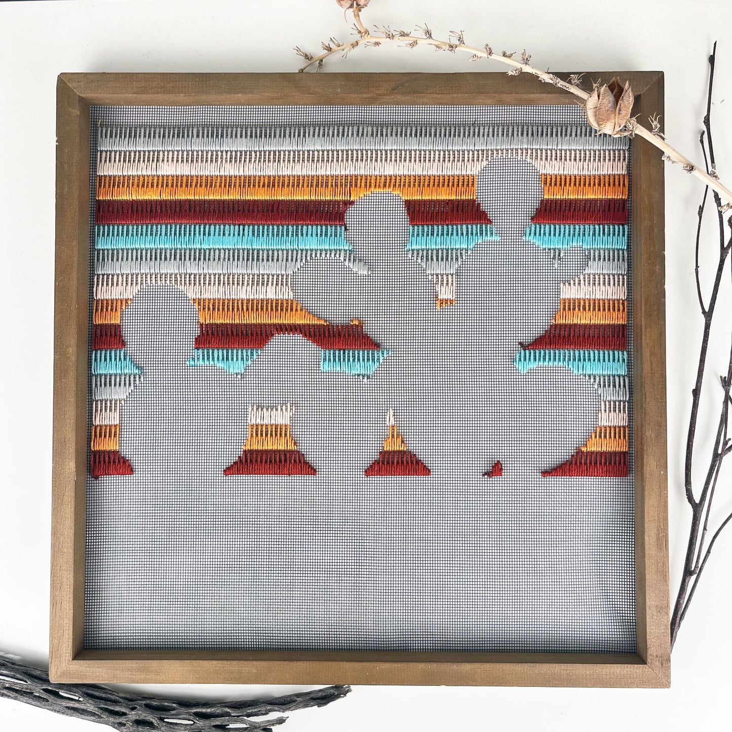 a piece of window screen hand stitched with rows of maroon orange peach grey and aqua blue stitches with an image in negative space of a prickly pear cactus, in a square wood frame, on a white counter, with dried yucca pods and cholla cactus around it
