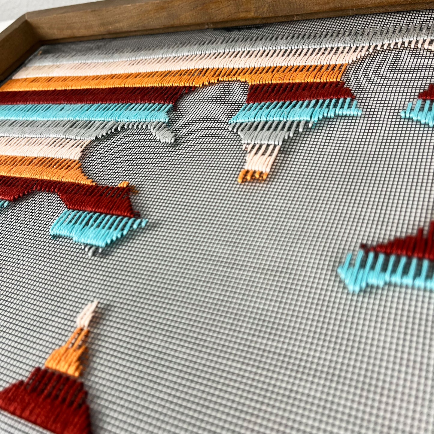 close up angled view of a piece of window screen hand stitched with rows of maroon orange peach grey and aqua blue stitches with an image in negative space of a prickly pear cactus, in a square wood frame, on a white counter