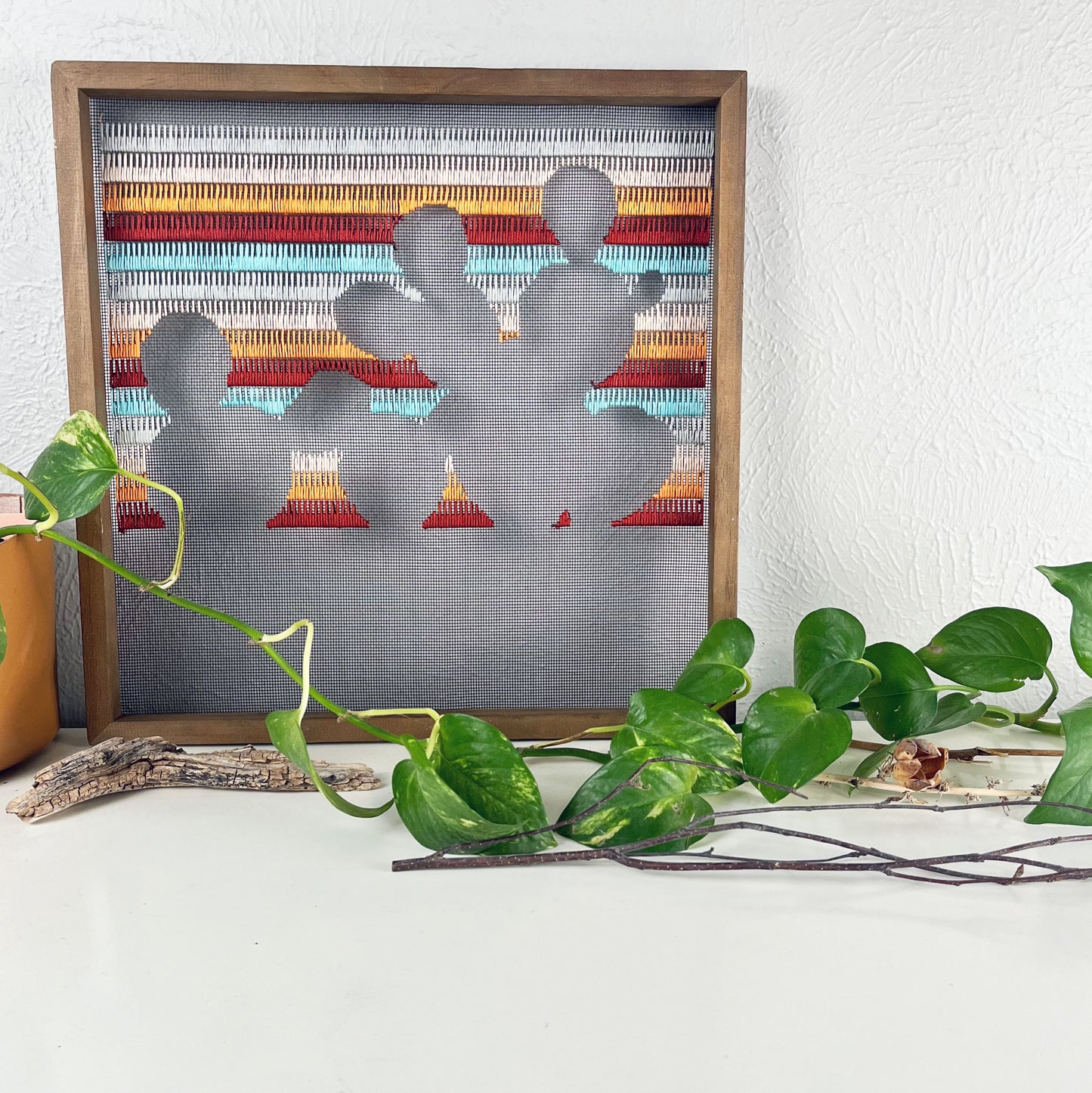 close up view of a piece of window screen hand stitched with rows of maroon orange peach grey and aqua blue stitches with an image in negative space of a prickly pear cactus, in a square wood frame, on a white counter, with a pothos vine around it