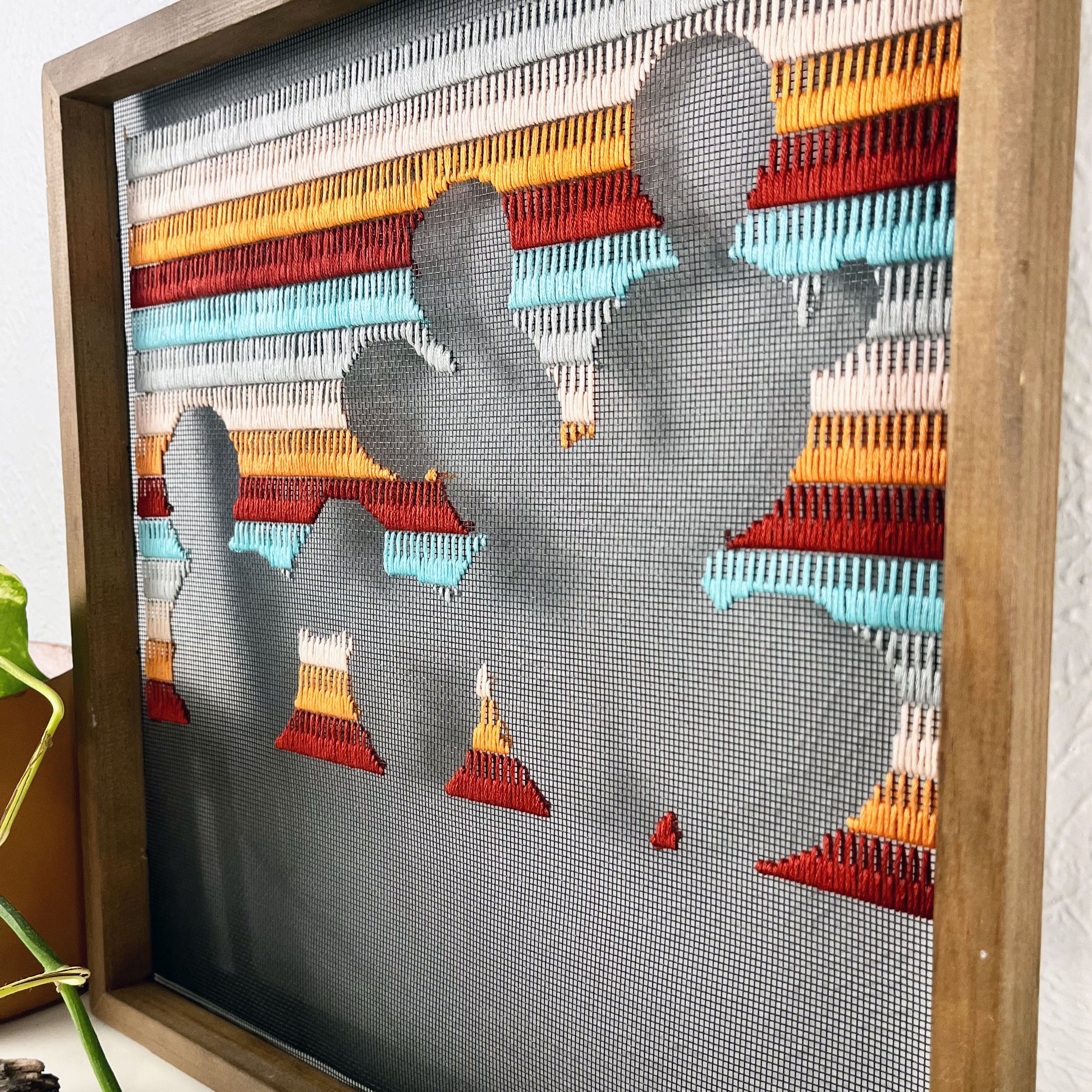close up angled view of a piece of window screen hand stitched with rows of maroon orange peach grey and aqua blue stitches with an image in negative space of a prickly pear cactus, in a square wood frame, on a white counter, with a pothos vine around it