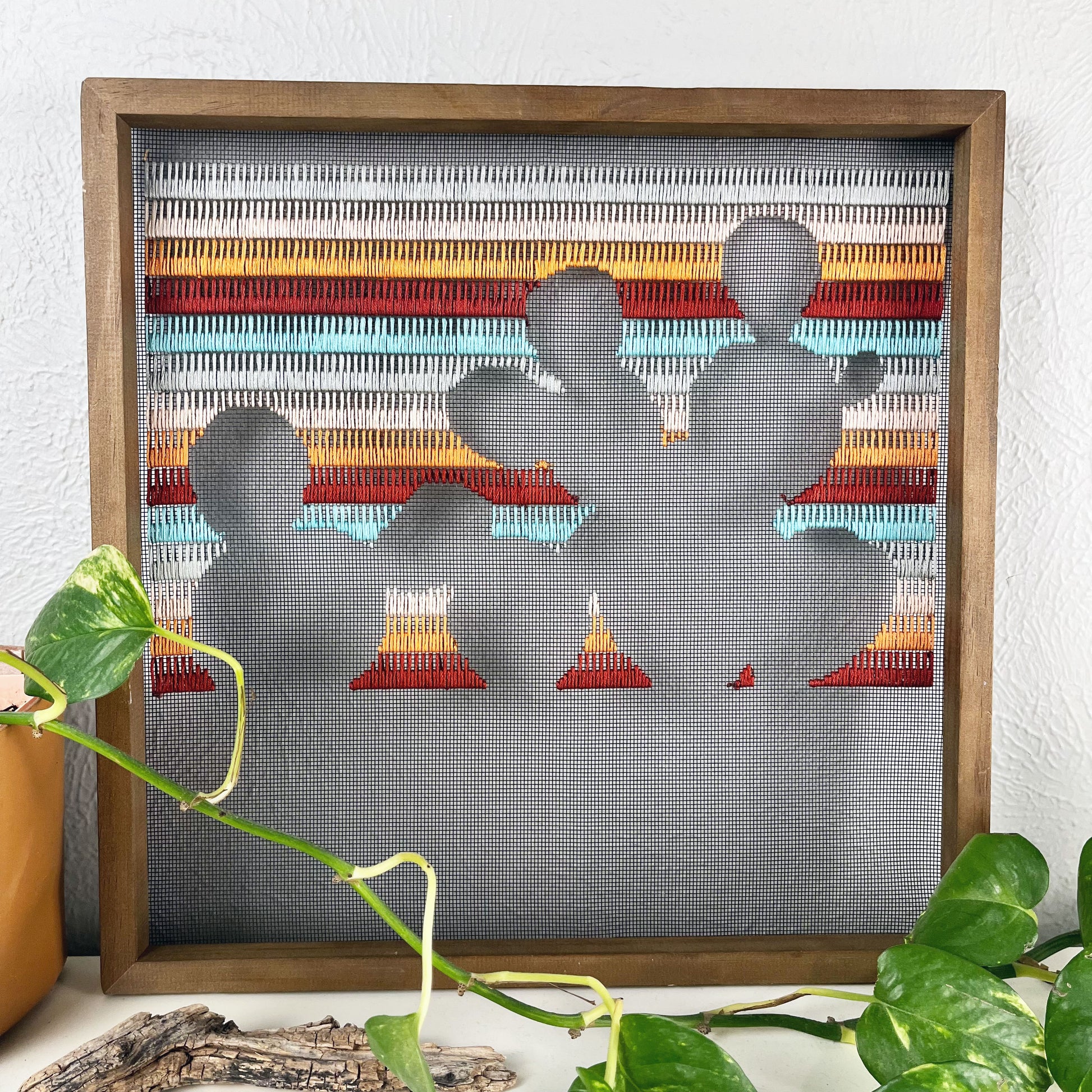 a piece of window screen hand stitched with rows of maroon orange peach grey and aqua blue stitches with an image in negative space of a prickly pear cactus, in a square wood frame, on a white counter, with a pothos vine around it