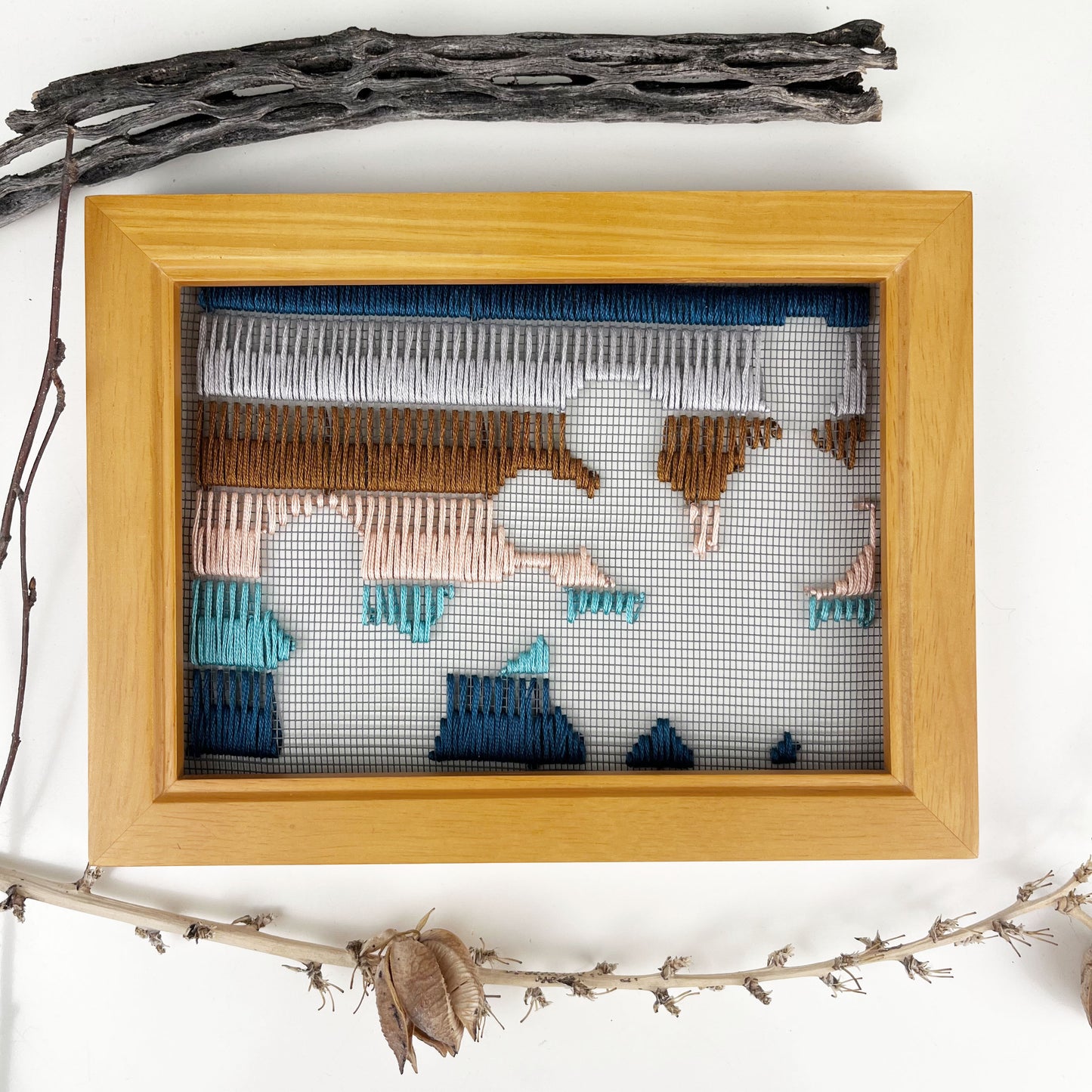 a piece of window screen hand stitched with rows of teal turquoise peach brown and grey stitches with an image in negative space of prickly pear cacti, in a brown wood frame, flat on a white counter, with dried cholla and yucca around it