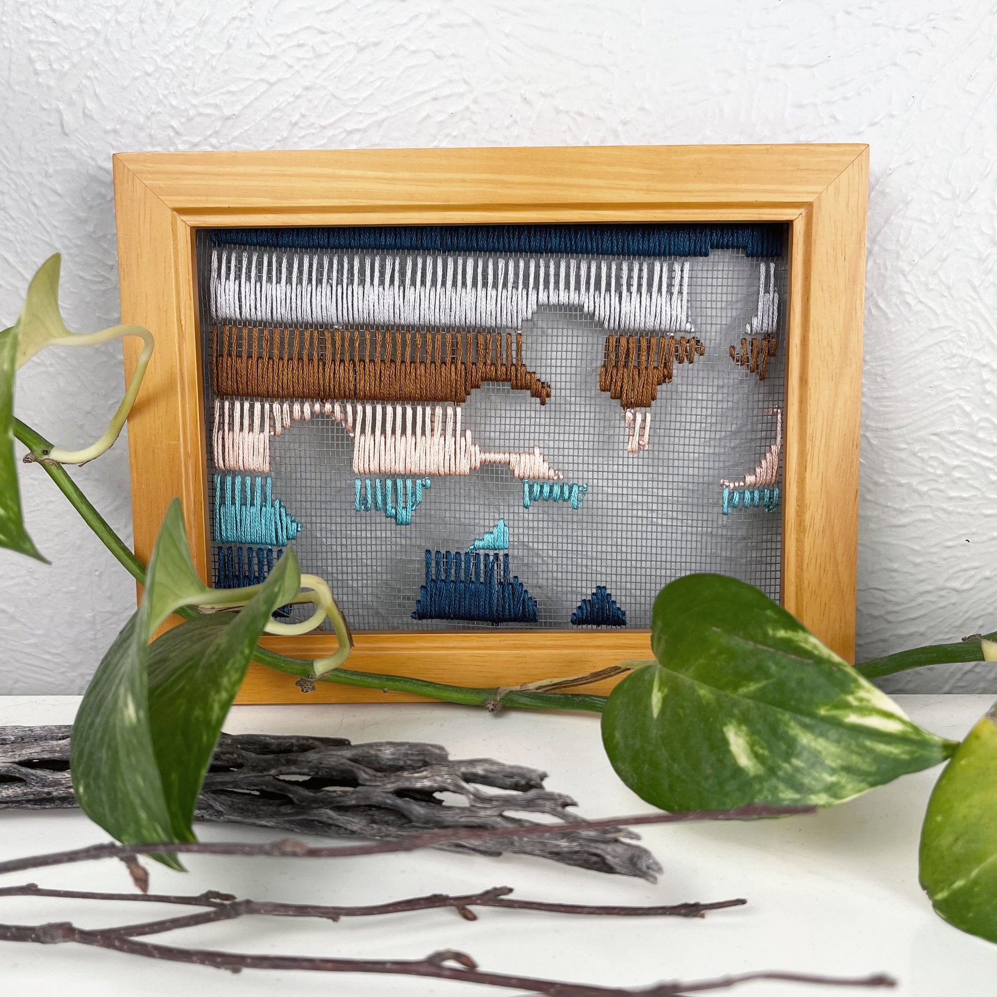 a piece of window screen hand stitched with rows of teal turquoise peach brown and grey stitches with an image in negative space of prickly pear cacti, in a brown wood frame, on a white counter, with a pothos vine around it