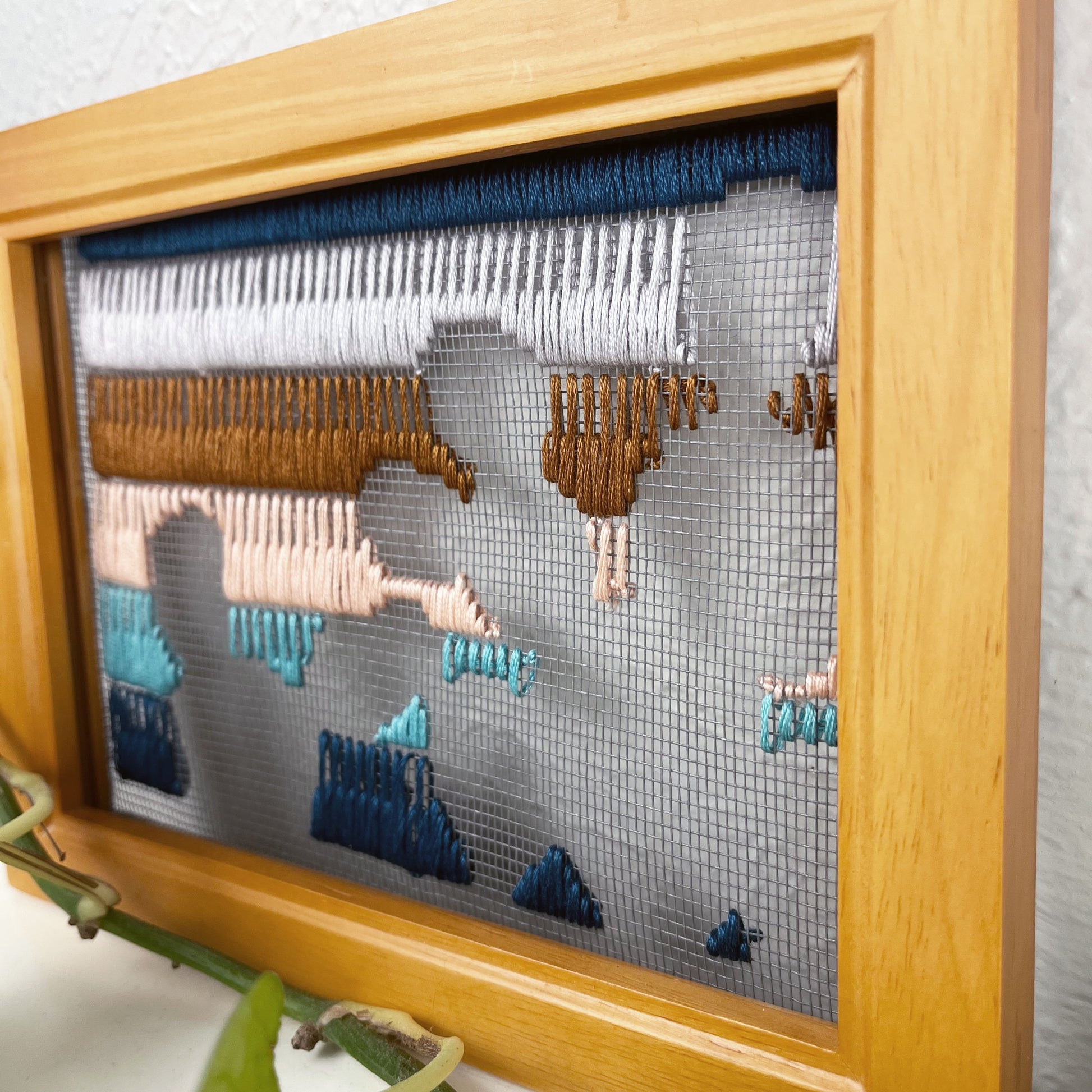 close up angled view of a piece of window screen hand stitched with rows of teal turquoise peach brown and grey stitches with an image in negative space of prickly pear cacti, in a brown wood frame, on a white counter, with a pothos vine around it