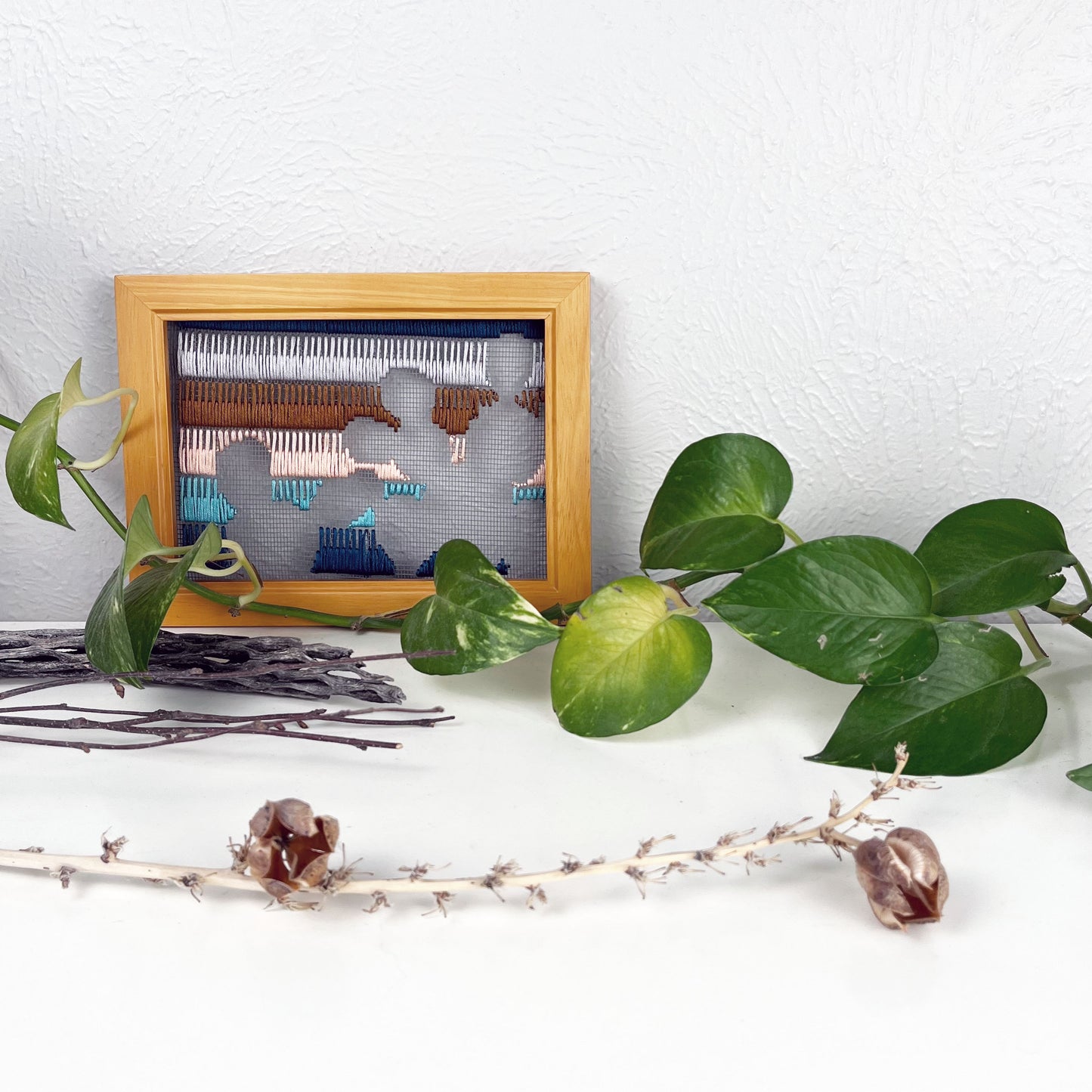 a piece of window screen hand stitched with rows of teal turquoise peach brown and grey stitches with an image in negative space of prickly pear cacti, in a brown wood frame, on a white counter, with a pothos vine around it