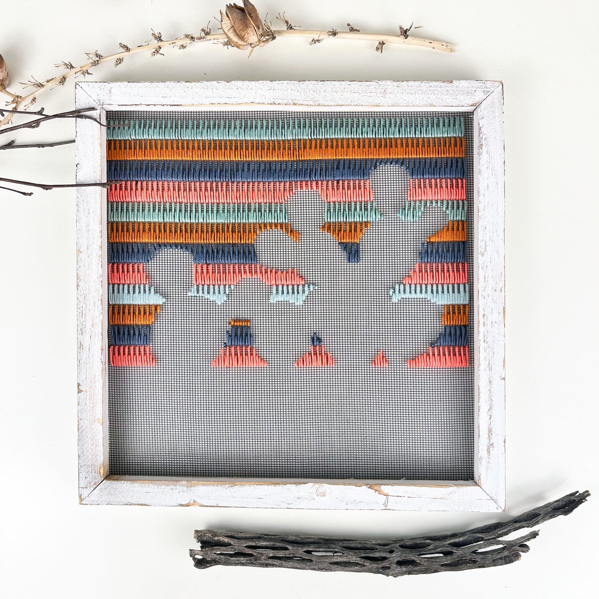 a piece of window screen hand stitched with rows of grey mustard mint green and neon pink stitches with an image in negative space of a prickly pear cactus, in a rustic white wood square frame, on a white counter, with dried yucca and cholla cactus around it