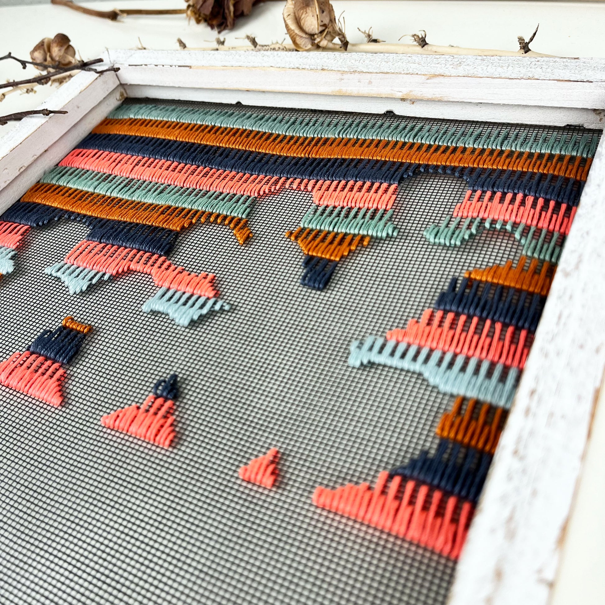 close up angled view of a piece of window screen hand stitched with rows of grey mustard mint green and neon pink stitches with an image in negative space of a prickly pear cactus, in a rustic white wood square frame, on a white counter, with dried yucca and cholla cactus around it