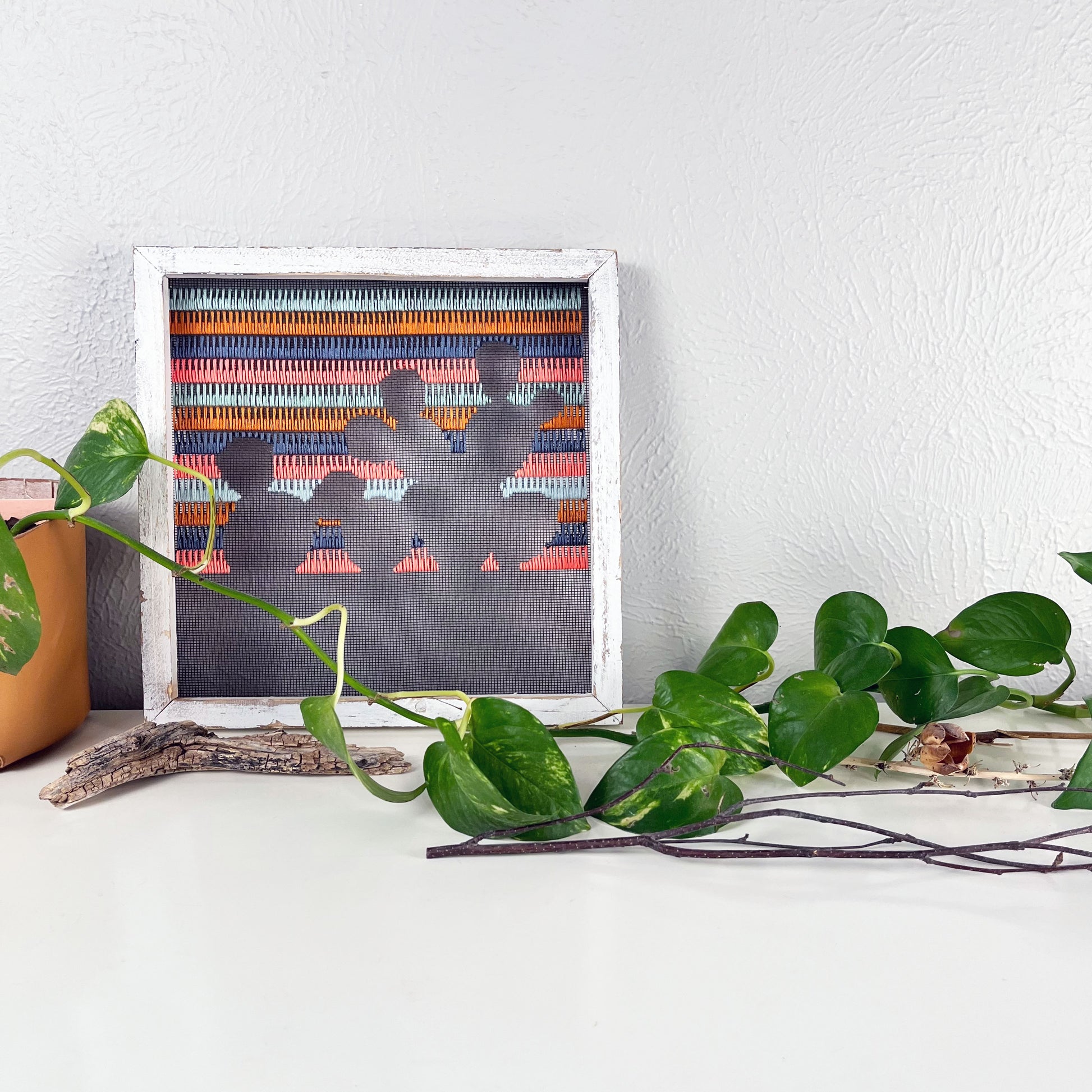 a piece of window screen hand stitched with rows of grey mustard mint green and neon pink stitches with an image in negative space of a prickly pear cactus, in a rustic white wood square frame, on a white counter, with a pothos plant and piece of found wood in front of it