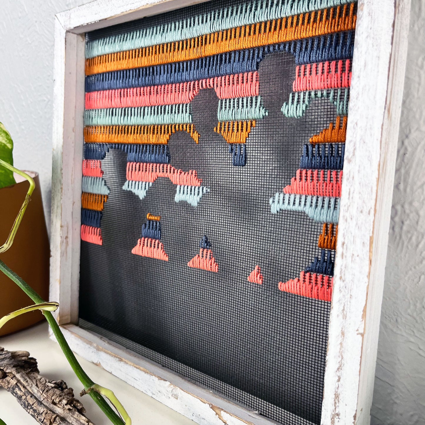 a close up angled view of a piece of window screen hand stitched with rows of grey mustard mint green and neon pink stitches with an image in negative space of a prickly pear cactus, in a rustic white wood square frame, on a white counter, with a pothos plant and piece of found wood in front of it