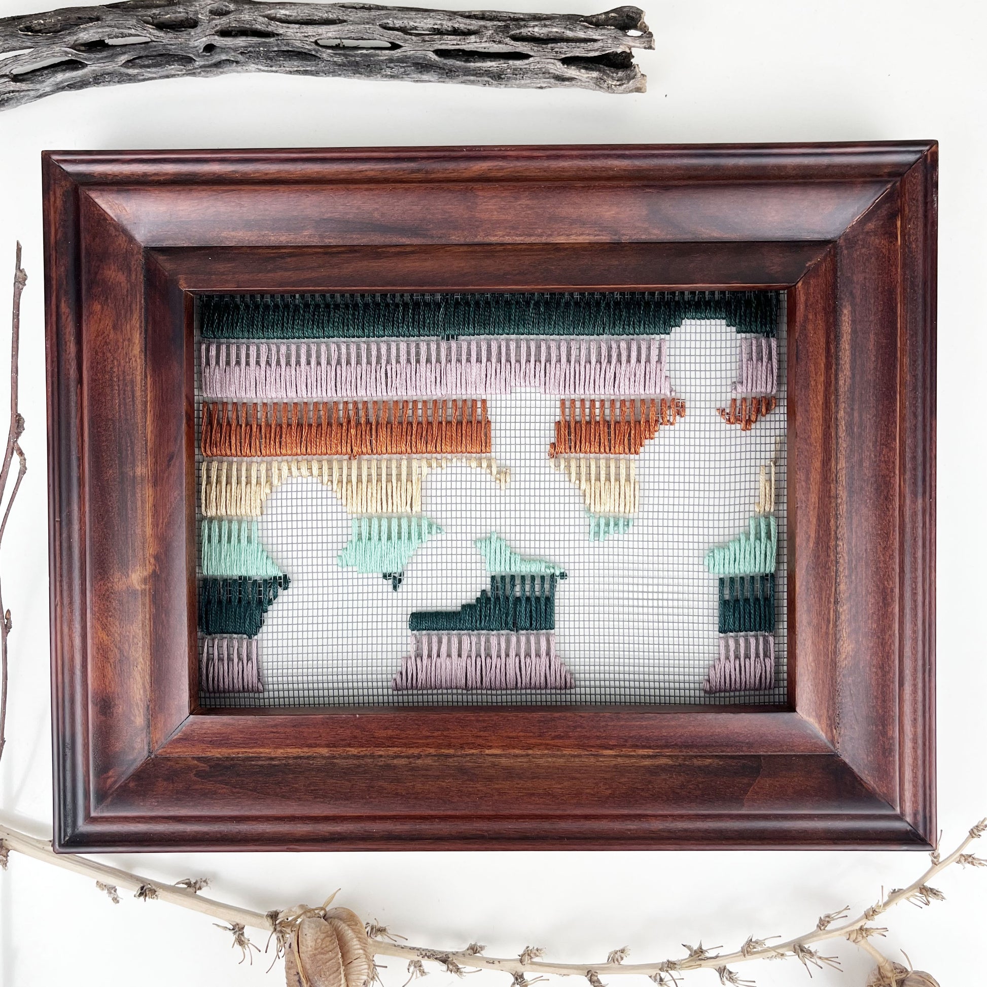 a piece of window screen hand stitched with rows of lavender forest green mint green peach and brown stitches with an image in negative space of prickly pear cacti, in a brown wood frame, flat on a white counter, with dried cholla and yucca around it