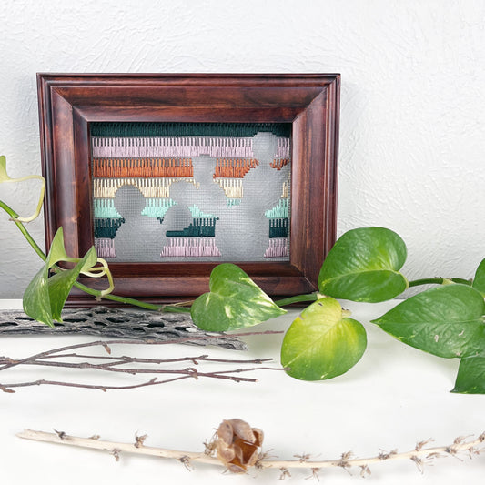 a piece of window screen hand stitched with rows of lavender forest green mint green peach and brown stitches with an image in negative space of prickly pear cacti, in a brown wood frame, on a white counter, with a pothos vine around it