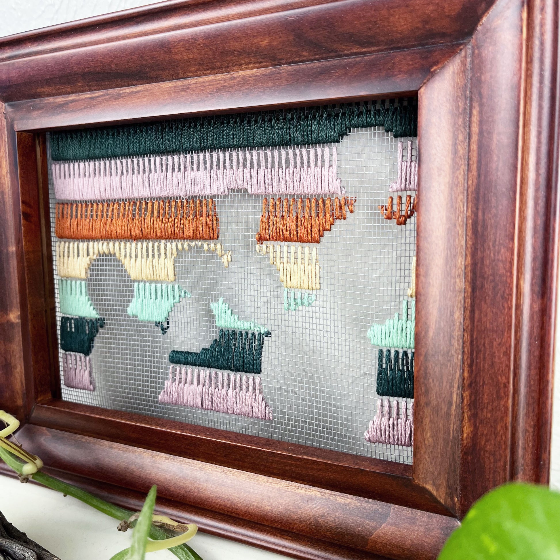 close up angled view of a piece of window screen hand stitched with rows of lavender forest green mint green peach and brown stitches with an image in negative space of prickly pear cacti, in a brown wood frame, on a white counter, with a pothos vine around it