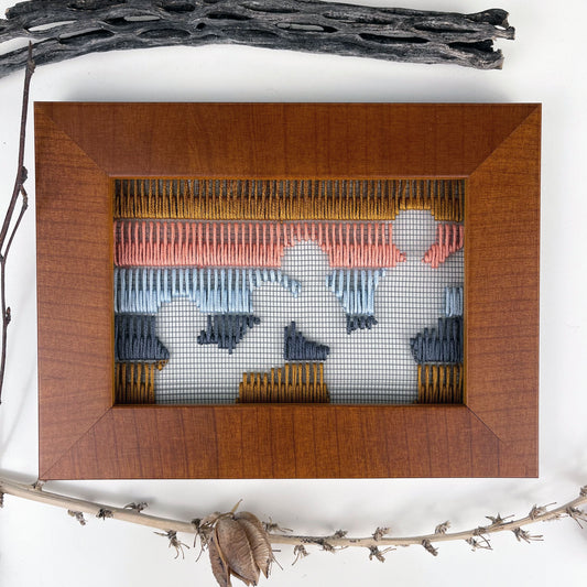 a piece of window screen hand stitched with rows of brown grey light blue coral and mustard stitches with an image in negative space of prickly pear cacti, in a brown wood frame, flat on a white counter, with dried cholla and yucca around it