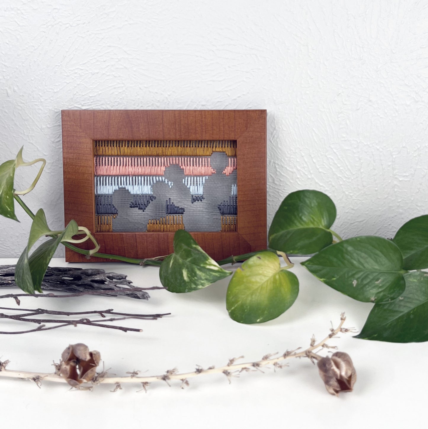 a piece of window screen hand stitched with rows of brown grey light blue coral and mustard stitches with an image in negative space of prickly pear cacti, in a brown wood frame, on a white counter, with a pothos plant dried cholla and yucca around it