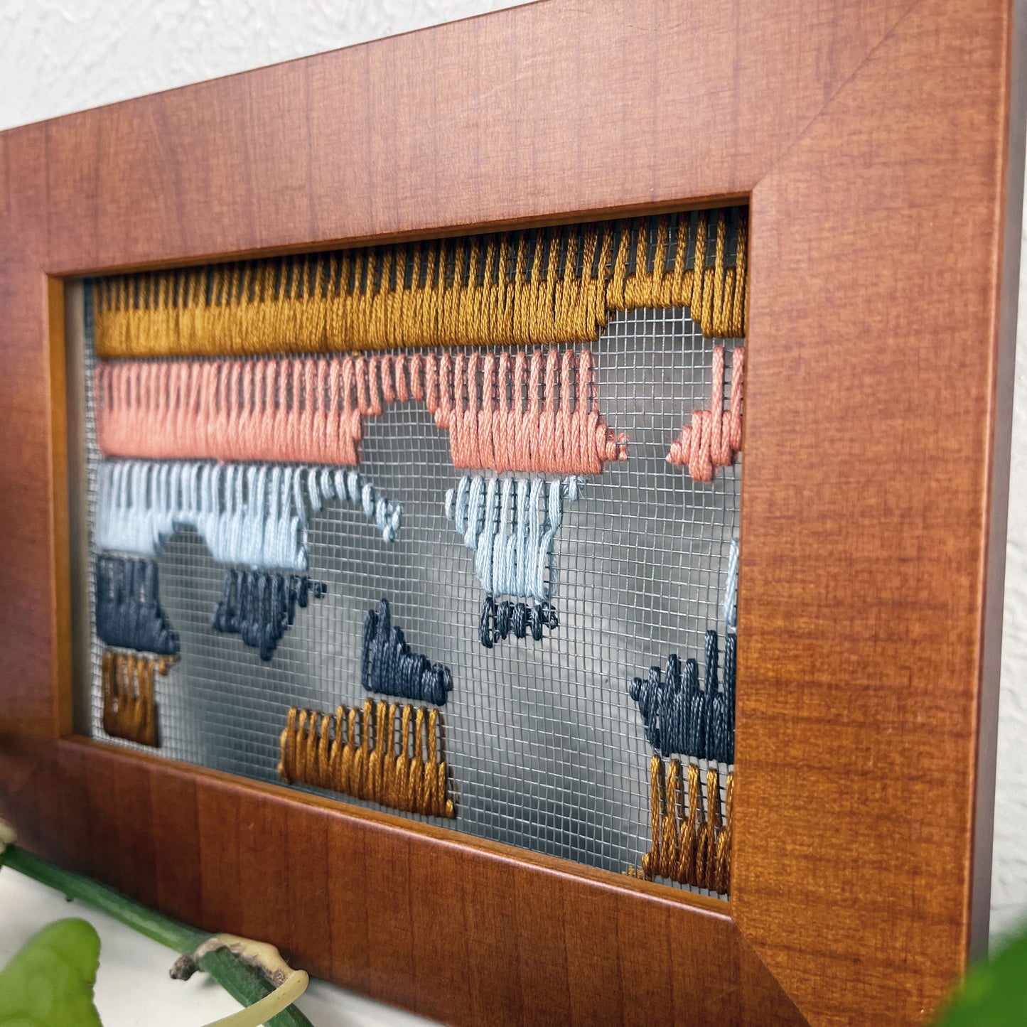 close up angled view of a piece of window screen hand stitched with rows of brown grey light blue coral and mustard stitches with an image in negative space of prickly pear cacti, in a brown wood frame, on a white counter, with a pothos plant dried cholla and yucca around it