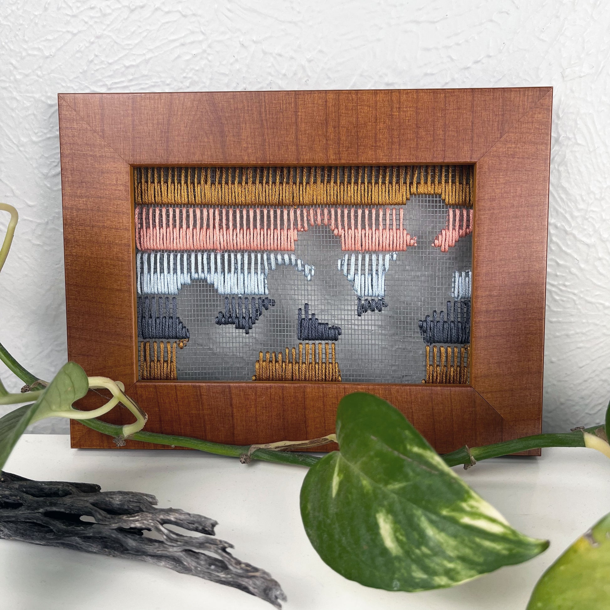 a piece of window screen hand stitched with rows of brown grey light blue coral and mustard stitches with an image in negative space of prickly pear cacti, in a brown wood frame, on a white counter, with a pothos plant dried cholla and yucca around it