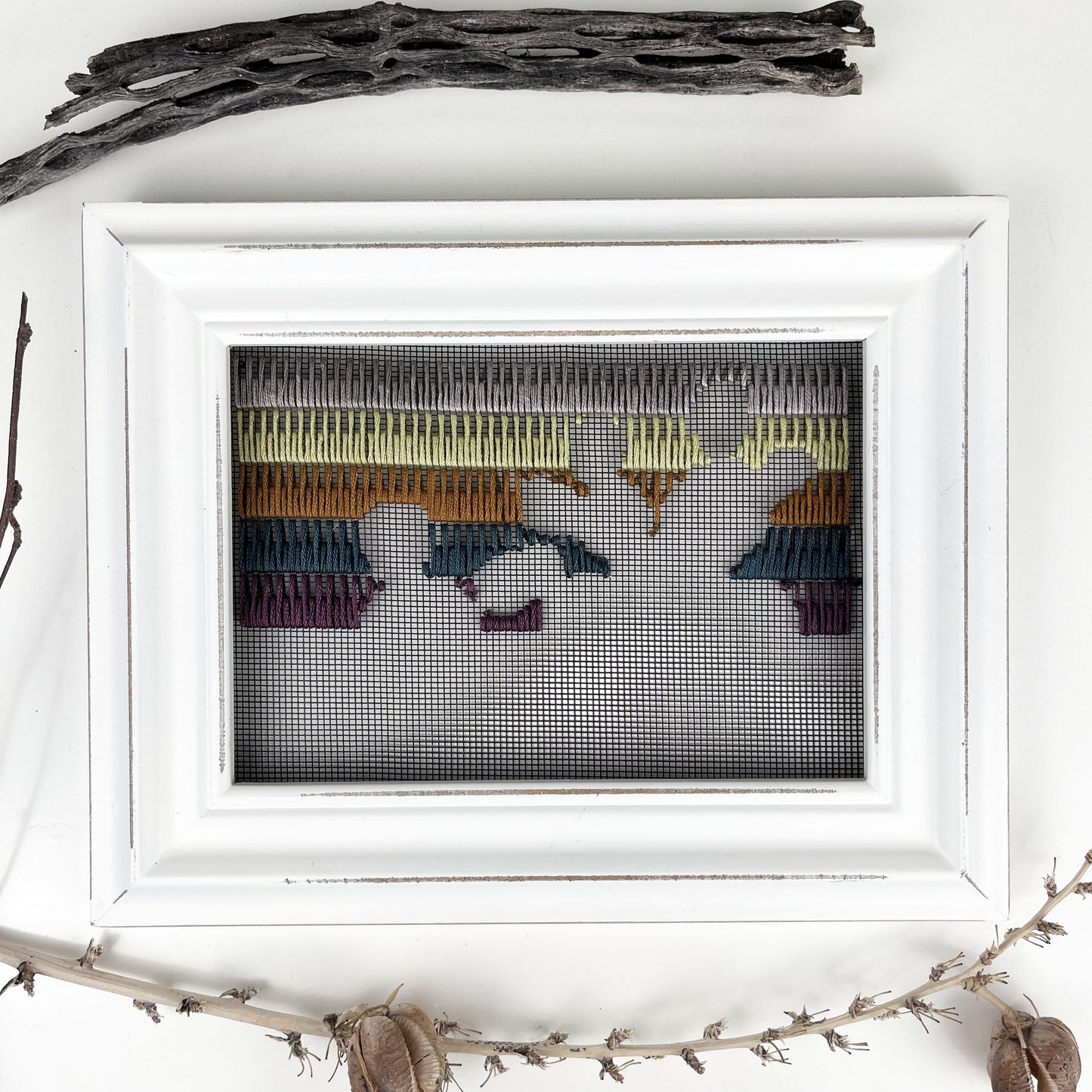 a piece of window screen hand stitched with rows of lavender chartreuse brown midnight blue and plum stitches with an image in negative space of prickly pear cacti, in a white wood frame, flat on a white counter, with dried cholla and yucca pods around it