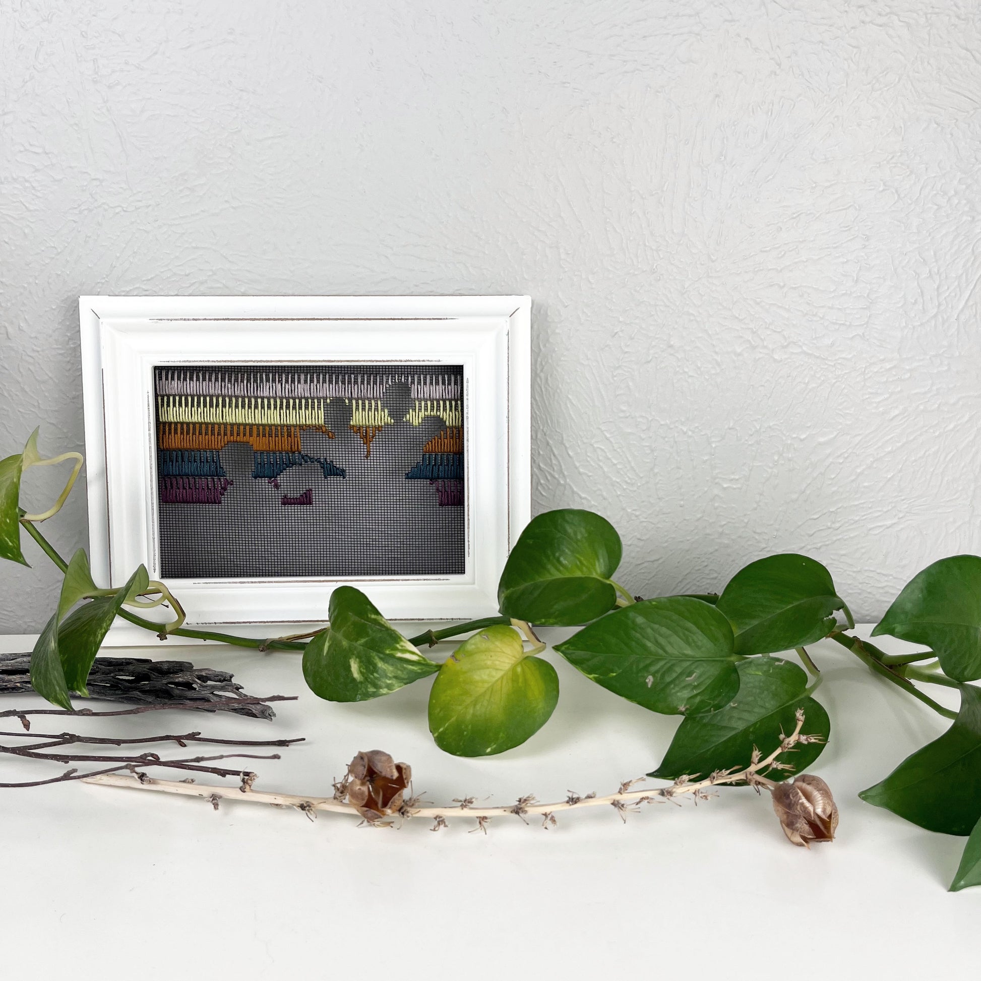 a piece of window screen hand stitched with rows of lavender chartreuse brown midnight blue and plum stitches with an image in negative space of prickly pear cacti, in a white wood frame, on a white counter, with a pothos vine around it